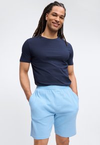 Άνδρας με μακριά dreadlocks που φοράει μπλε ναυτικό t-shirt και ανοιχτό μπλε υφασμάτινο σορτς με τα χέρια στις τσέπες, χαμογελώντας, σε απλό φόντο.