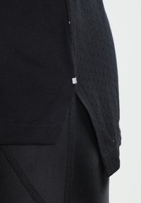 Gros plan d’une tenue de sport noire montrant des tissus superposés avec une petite fente latérale et un matériau perforé texturé.