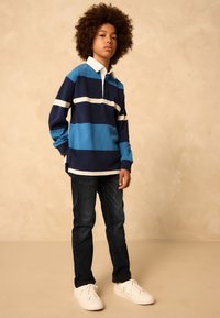 Mėlynas ir tamsiai mėlynas dryžuotas ilgomis rankovėmis polo marškinėlis su baltu apykakle, derinamas su tamsios spalvos džinsais ir baltais sportbačiais. Paprastas, kasdienis dizainas.