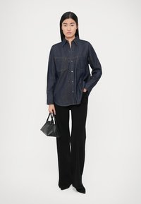 Camisa de ganga escura com botões e dois bolsos no peito, combinada com calças pretas de cintura alta. Acessórios incluem uma pequena mala preta.