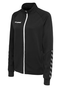 Chaqueta negra con cremallera y un tejido texturizado, cremallera blanca y un patrón en chevron en blanco y negro en las mangas. Presenta un cuello alto.