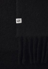 Tissu en laine noire avec une surface douce et texturée. Présente une petite étiquette blanche avec un logo noir. Comprend des détails plissés sur le bord.