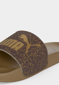 Καφέ σανδάλι Puma slide με λουράκι με λεοπάρ μοτίβο και χρυσλό λογότυπο Puma στην κορυφή σε απλό ανοιχτό φόντο.