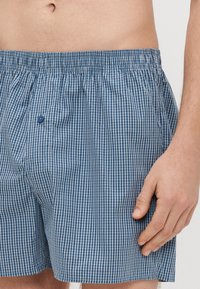 Modri in beli karo kratke hlačke z gumijastim pasom, s spodnjim zapiranjem na gumb in sproščenim krojem. Tkanina je videti lahka in zračna.