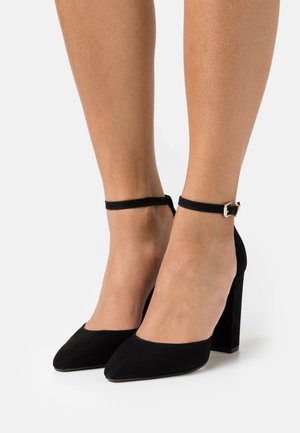 Escarpins en daim noir avec un design à bout pointu, bride de cheville avec une boucle dorée et un talon carré. Texture lisse, style classique.