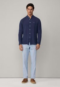 Camicia azzurra di lino con bottoni e colletto alla cinese, abbinata a pantaloni a righe blu chiaro e scarpe in suede marroni.