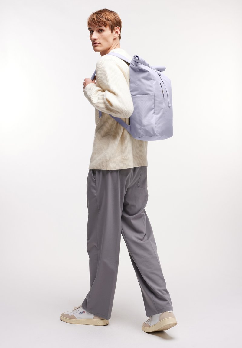 Svetlefialový batoh s rolovacím vrškem a zipovým uzávěrem, vyrobený z odolné látky. Nesený na jednom rameni, v kombinaci se krémovým svetr a šedými kalhotami.