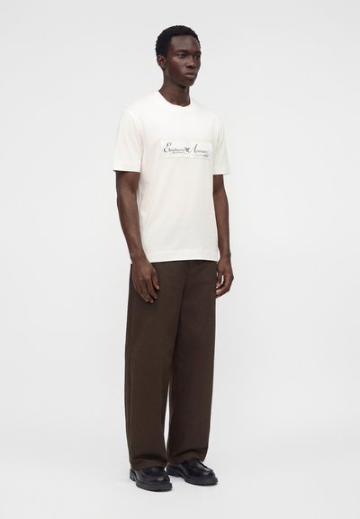 Άνδρας που φοράει λευκό t-shirt Emporio Armani, φαρδιά καφέ παντελόνια και μαύρα παπούτσια, στέκεται μπροστά σε ένα απλό φωτεινό φόντο.
