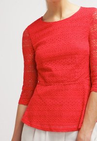Top en dentelle rouge avec des manches trois-quarts, présentant un motif zigzag texturé et un ourlet peplum. Design léger et ajusté.