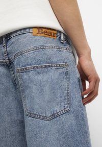 Primer plano de una persona que lleva puesto unos pantalones vaqueros de mezclilla azul claro con un gran bolsillo trasero y una etiqueta de marca de cuero marrón en la cinturilla.