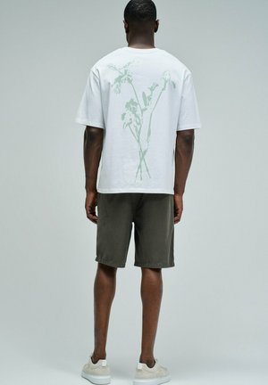 Hombre con camiseta blanca con un tenue estampado floral verde en la espalda, pantalones cortos oscuros y zapatillas de color claro, de pie frente a un fondo gris liso.