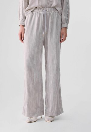 Donna che indossa pantaloni larghi grigio chiaro con coulisse in vita e tacchi bianchi aperti sulle punte su uno sfondo semplice.