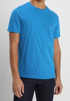 Mann trägt ein schlichtes, hellblaues Kurzarm-T-Shirt und dunkelblaue Jeans, steht vor einem hellgrauen Hintergrund.