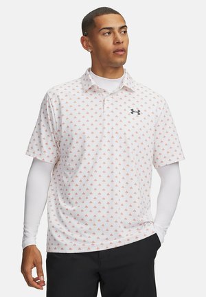 Mann trägt ein weißes kurzärmeliges Poloshirt mit kleinen orangefarbenen Mustern über einem weißen langärmeligen Hemd und schwarzen Hosen, schaut zur Seite.