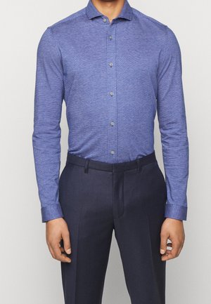 Blue long-sleeve shirt in a textured fabric, featuring a button-down collar and front buttons, paired with dark trousers.