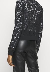 Mustat pitsijakku, jossa on kukkakuosi ja ribbineulokset, päällä mustat keinonahkahousut. Läpinäkyvä kangas paljastaa alla olevan kerroksen.