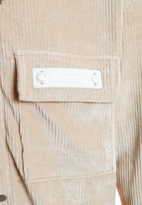 Camisa de sarga beige con un bolsillo en el pecho y etiqueta blanca. Presenta una textura de canalé vertical y se cierra con botones de metal.