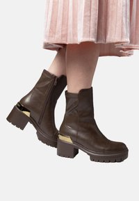 Botas de cuero marrón hasta el tobillo con cierre de cremallera, tacón cuadrado y un acento dorado en el tacón. Suela texturizada para un mejor agarre y estabilidad.