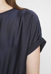 Gros plan d'une personne portant une chemise à manches courtes bleu marine avec un revers sur la manche, sur un fond uni.