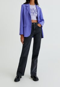 Donna che indossa un blazer viola, un top grafico corto, jeans neri a vita alta e stivali neri chunky, in piedi con una mano in tasca.