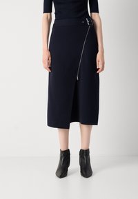 Jupe midi bleu marine avec une taille structurée, détail de fermeture éclair diagonal et tissu lisse et texturé. Associée à des bottines noires à talons pointus.