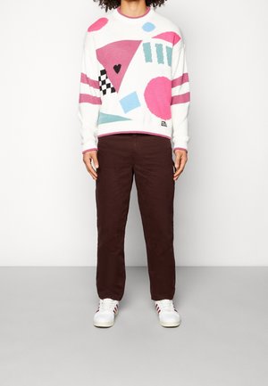 Osoba oblečená v bílém svetru s růžovými, modrými a černými geometrickými tvary, tmavě hnědých kalhotách a bílých teniskách s červenými pruhy.