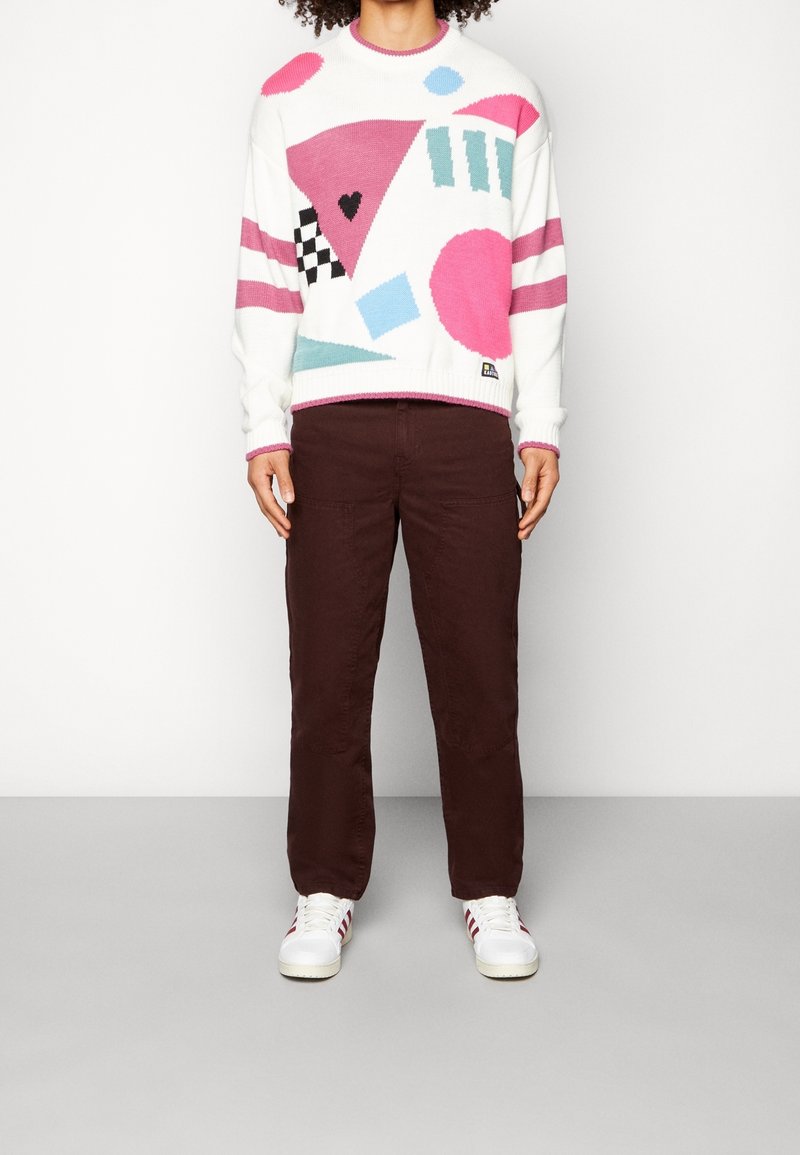 Persona con un suéter blanco con formas geométricas en rosa, azul y negro, pantalones marrón oscuro y zapatillas blancas con rayas rojas.