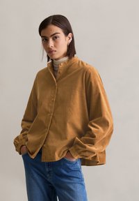 Chemise en velours côtelé marron avec un col haut et un devant à boutons, présentant des poignets larges et froncés. Portée sur un turtleneck clair et un jean bleu.