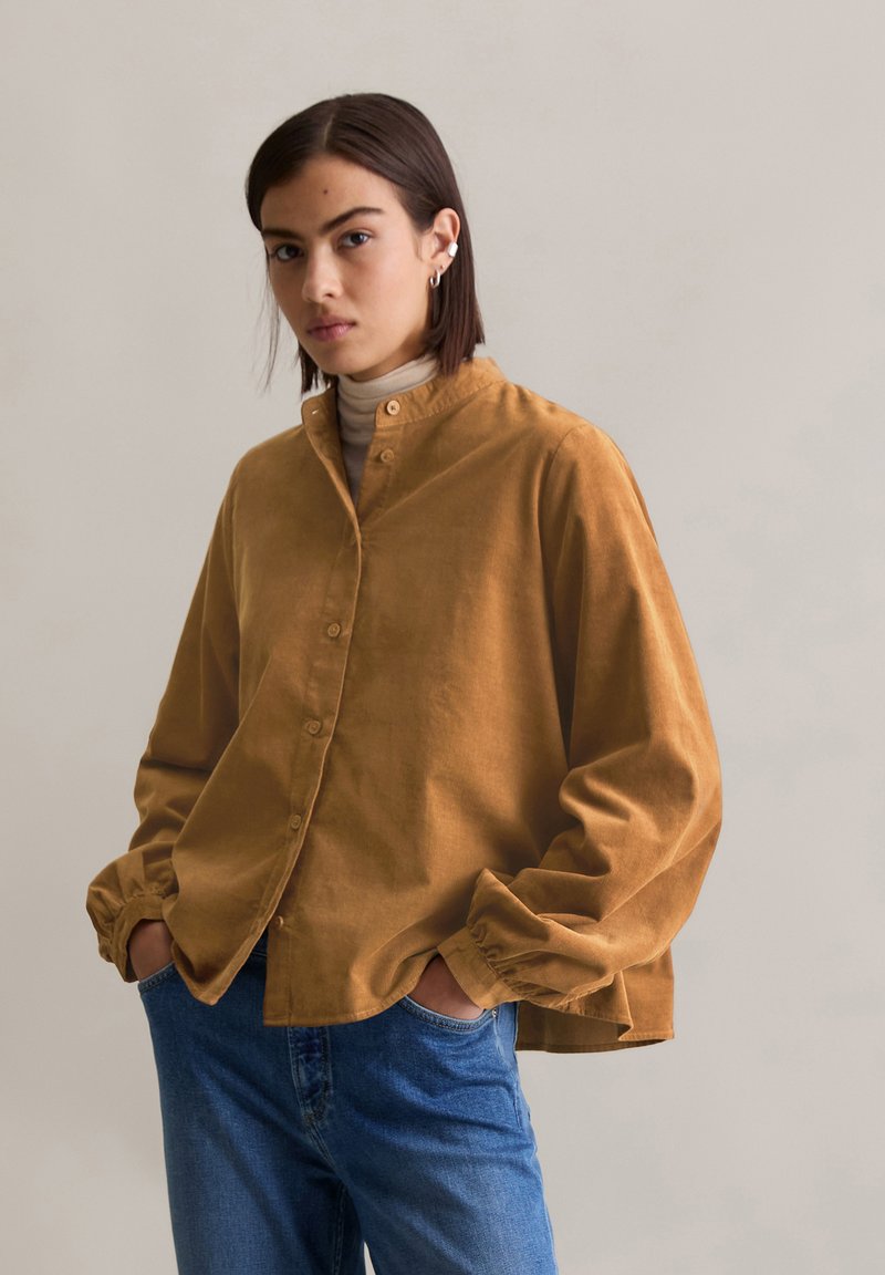 Chemise en velours côtelé marron avec un col haut et un devant à boutons, présentant des poignets larges et froncés. Portée sur un turtleneck clair et un jean bleu.