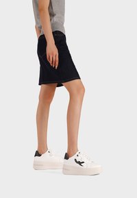 Persona che indossa una gonna di jeans scura fino al ginocchio e scarpe sportive bianche con dettagli neri, in piedi di profilo su uno sfondo semplice.