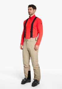 Beige Skihosen mit schwarzen Akzenten, verstellbaren Trägern und einer glatten Textur, kombiniert mit einem enganliegenden roten Langarmshirt.