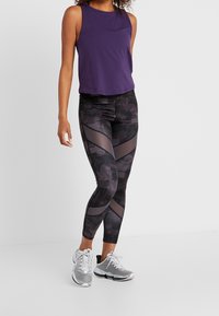 Fialový bezrukávový top v kombinaci s černými legínami s texturovaným vzorem a síťovými prvky. Šedé sportovní boty dokončují celý outfit.