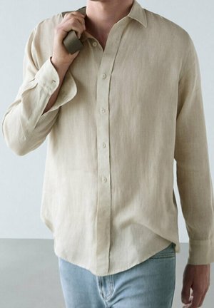 Mężczyzna w beżowej koszuli z długim rękawem i jasnoniebieskich dżinsach, trzymający zielony pasek na ramieniu.