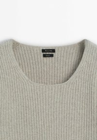Pull en laine côtelée beige clair avec encolure arrondie et étiquette noire indiquant la marque et les détails du matériau.