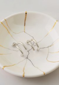 Silverörhänge med en böjd design, utställda på en vit tallrik med guld- och svart marmorering. Slät textur med geometriska accenter.