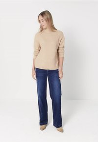 Beżowy sweter z dzianiny o teksturowanym wzorze i ściągaczami, w zestawieniu z ciemnoniebieskimi spodniami o szerokich nogawkach oraz beżowymi butami na ostrej końcówce.