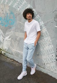Hvid kortærmet T-shirt med sort logo, lyseblå snorede bukser og hvide sportssko med pink accenter, sat mod en graffiti-væg.
