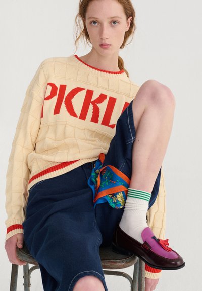 Jeune personne à la peau claire et aux cheveux roux portant un pull crème avec l'inscription « PICKLE » en rouge, un pantalon ample foncé, des chaussettes blanches et des mocassins violets, assise sur une chaise.