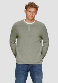  Lichtgroene gebreide trui met een gestructureerd patroon, ronde hals en een placket met drie knopen. Gecombineerd met blauwe jeans, met ontspannen armen langs de zijkanten.
