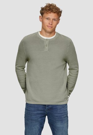 Pull en maille vert clair avec un motif texturé, col rond et patte de boutonnage à trois boutons. Associé à un jean bleu, les bras relaxés le long des côtés.