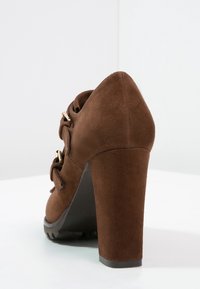 Scarpa con tacco alto in suede marrone con punta arrotondata, caratterizzata da un fiocco decorativo e dettagli in acciaio inossidabile dorato, e un tacco texturizzato per una migliore aderenza.
