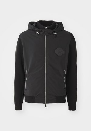 Chaqueta negra con cremallera en el frente, capucha, puños acanalados y cremalleras en los bolsillos. Presenta un logo de parche texturizado en el pecho y mangas de punto suave.