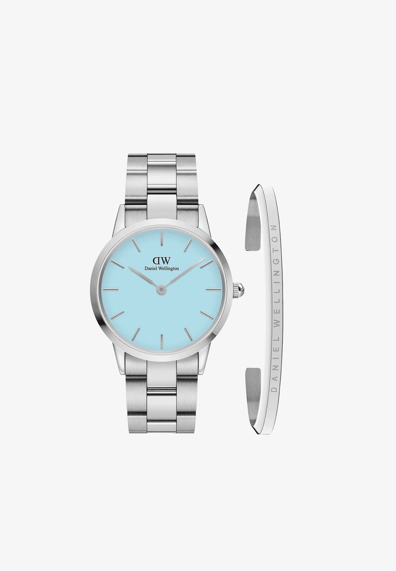 Hõbedane roostevabast terasest kell ümmarguse korpuse, taevasinise näo, kitsaste tundemarkerite ja vastava metallist käevõruga. Sisaldab eemaldatavat rihma.