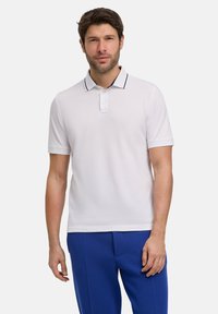 Męska koszulka polo z krótkim rękawem w kolorze białym, z dwu guzikowym kołnierzykiem i granatowymi akcentami. Gładka tkanina, luźny krój. Nosi wystylizowane z niebieskimi spodniami.