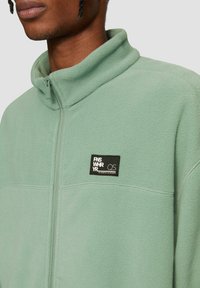 Fleecejacke in Hellgrün, mit Reißverschluss am Hals, weicher Textur, mit einem schwarzen Logopatch auf der linken Brust und flachen Nahtdetails.