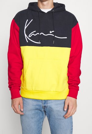 Person trägt einen Color-Block-Hoodie mit schwarzem Oberteil, gelber Brustpartie, roten Ärmeln und weißem Schriftzug, kombiniert mit hellblauen Jeans.