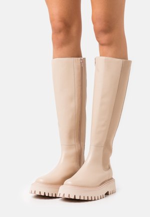 Bottes en cuir beige jusqu'aux genoux avec une semelle à plateforme épaisse, fermeture éclair sur le côté et texture lisse ; arborant un design minimal et des panneaux latéraux contrastants.