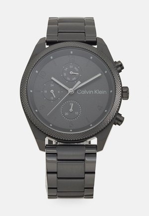 Črno nerjaveče jekleno uro Calvin Klein z obrobo z teksturo, sivo številčnico, tremi sub-številčnicami in povezanimi zapestnicami. Opremljena je s srebrnimi kazalci in logotipom.