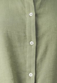 Camisa de lino verde claro con cierre de botones y botones blancos. La textura es suave y un poco áspera, mostrando fibras naturales.