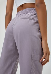 Pantalon cargo lilas avec taille élastique, poche latérale et texture en coton lisse. Présente un détail de poche arrière cousue.